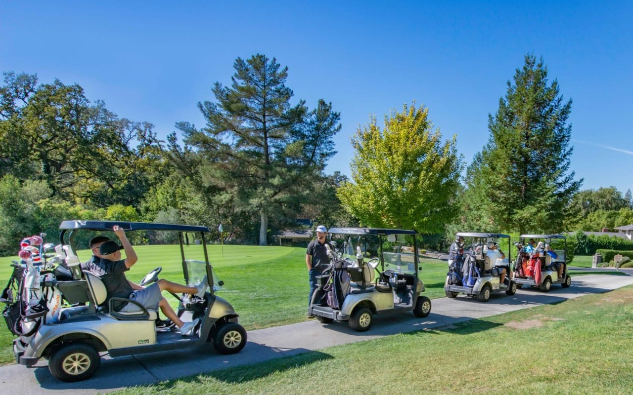  A bright, sunny day at a golf course features a line of four golf carts on a paved path. 
