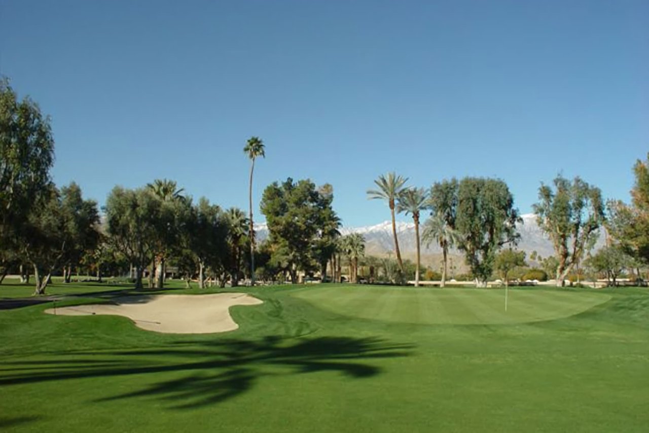 A golf course green with a sand trap and flag, and with palm trees and other mature trees.