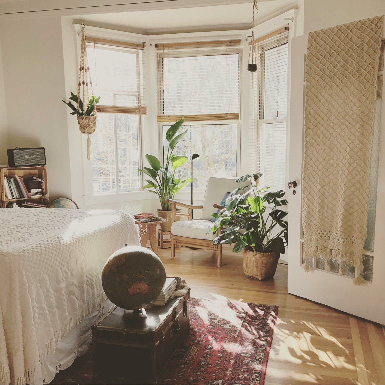 White bedroom with plants a red rug armchair wooden chest and bay window