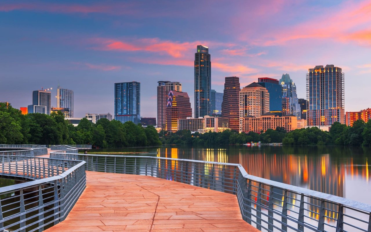 A wooden boardwalk with metal railings over a lake in a city at night, with distant skyscrapers and trees on the shore.