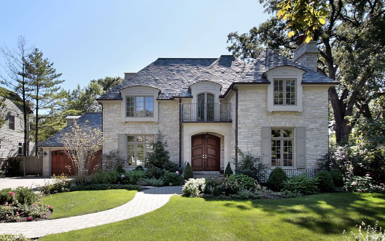 Symmetrical two-story brick house in a wooded area with slate roof, chimney, covered porch, and front lawn.