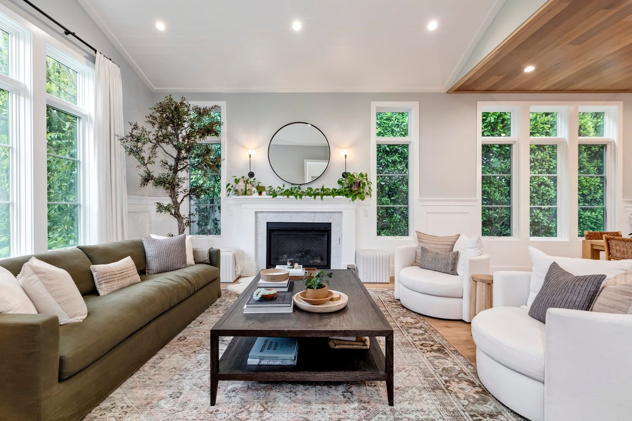  A well-decorated and bright living room features a long, olive-green sofa on the left and two white armchairs on the right, arranged around a dark wooden coffee table with a light-colored tray holding decorative items and books. 
