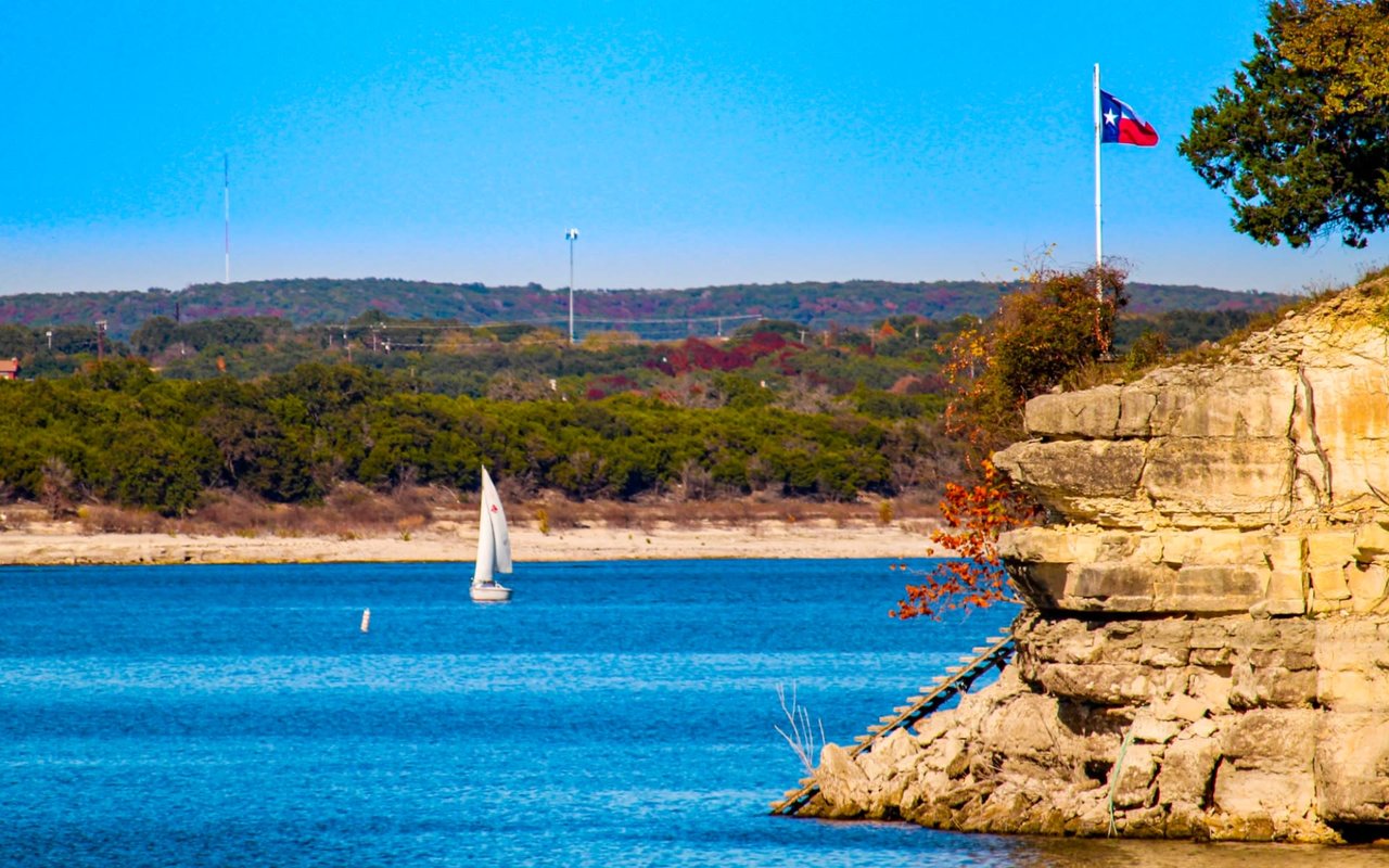 A white sailboat glides on the serene Lake Travis, surrounded by green hills, trees, houses, and docks under a cloudless sky.