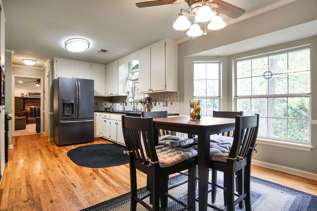 The kitchen has white cabinets, black countertops, and wooden flooring. The dining area includes a table with chairs and is well-lit by a large window.