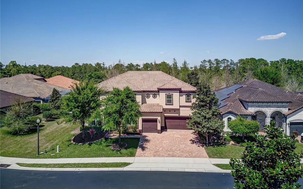 Two-story stucco home with a tan tile roof, brick-paver driveway, and mature landscaping.