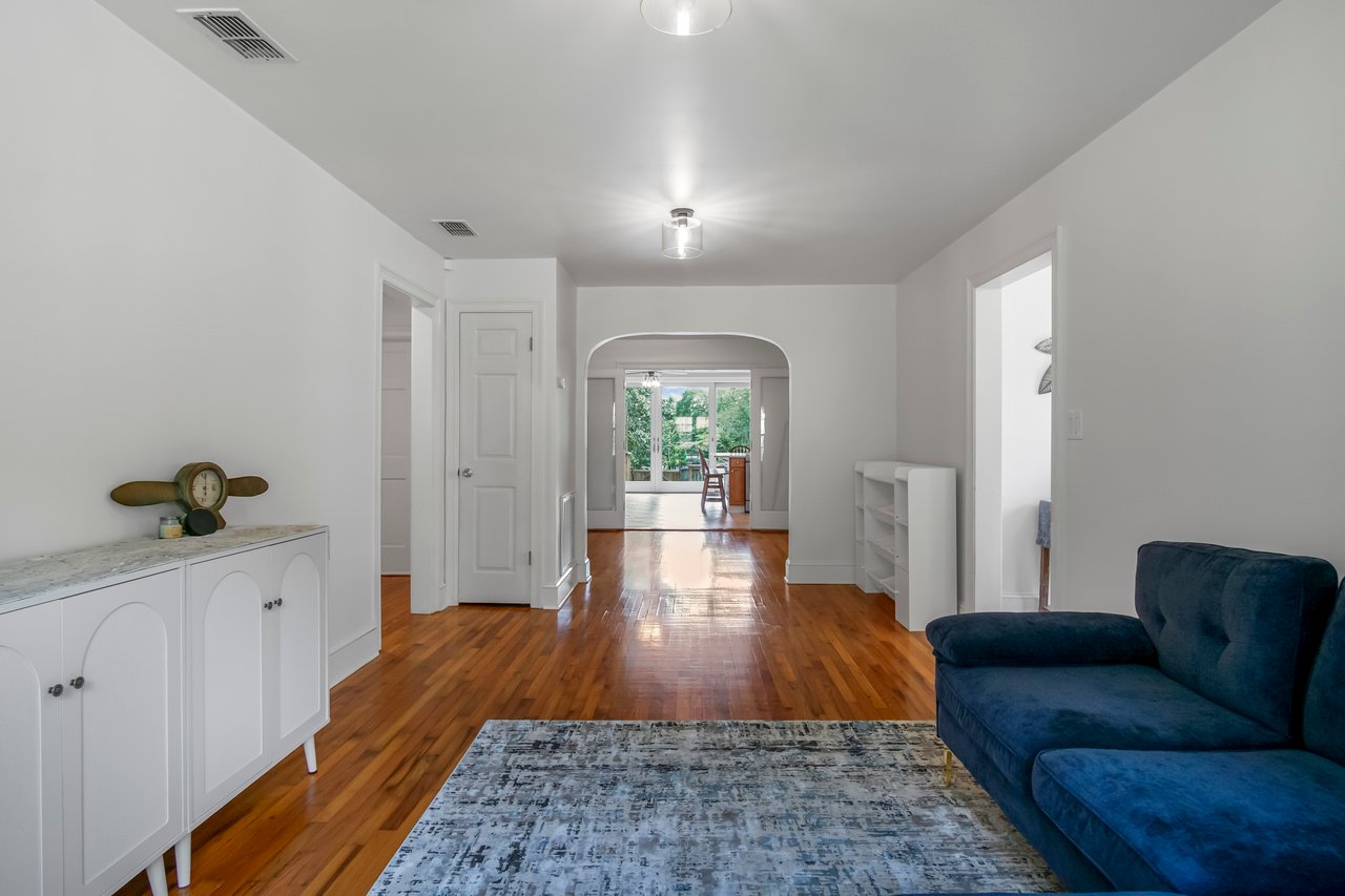 A bright, minimalist living room with white walls, wooden floors, a blue sofa, a white sideboard, and a rug. Archway leads to a sunlit area. Calm ambiance.