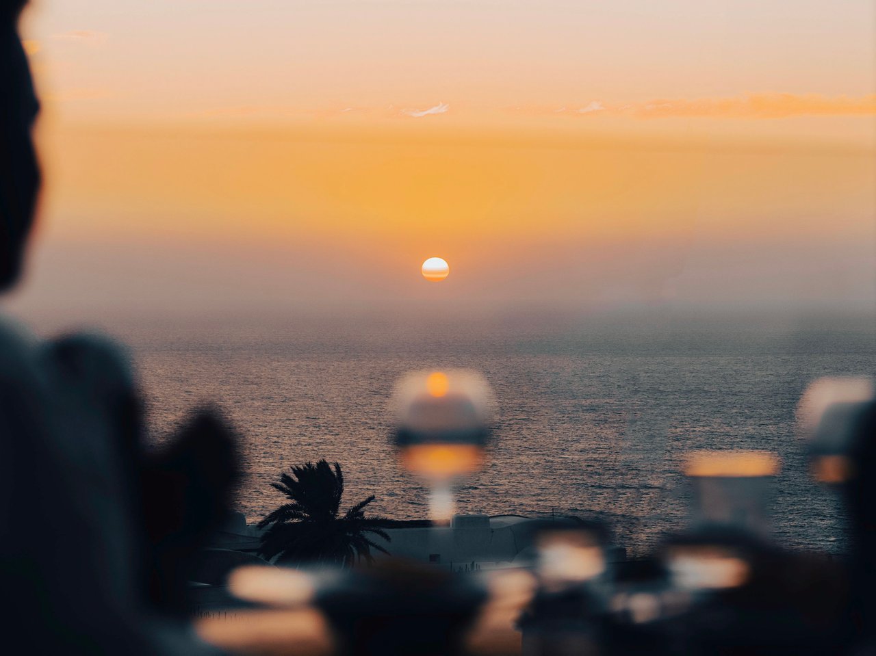 Orange sun setting over the ocean, framed by blurred foreground lights and silhouettes.
