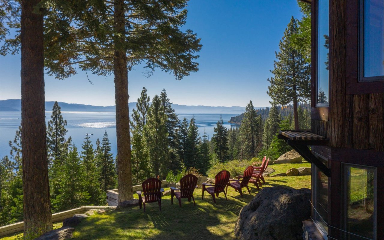 Lake Tahoe view from a lodge terrace, framed by pine trees and Adirondack chairs.