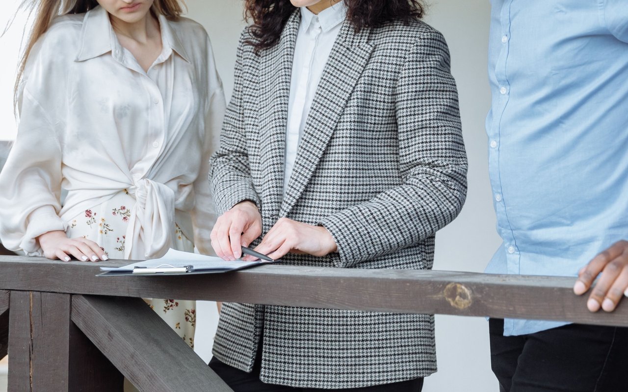 Three people review paperwork at a wooden railing as one writes on a clipboard.