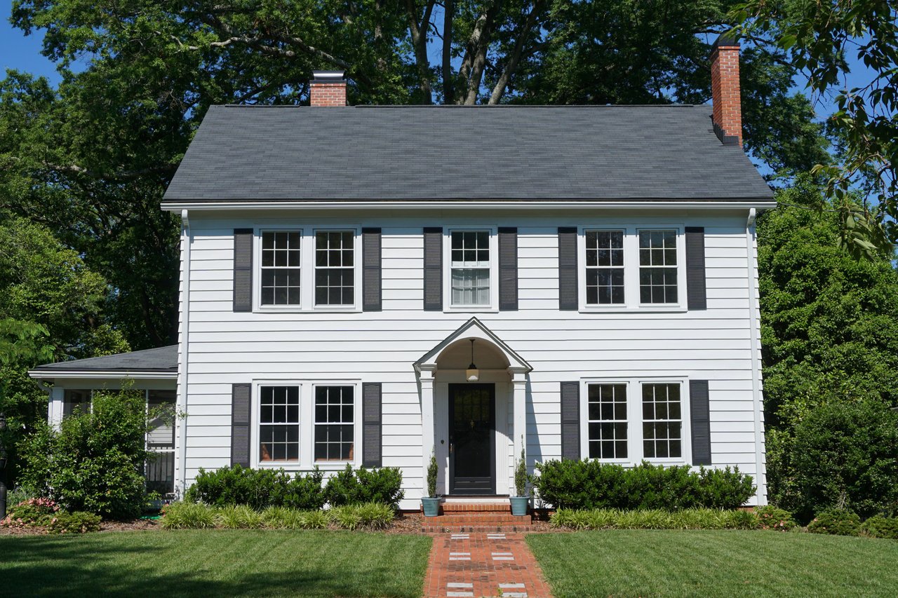 White two-story house with dark shutters and a brick walkway to its arched front entry.
