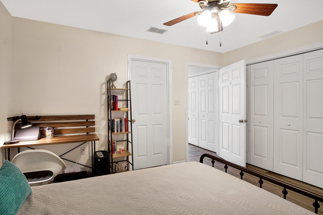 Bright bedroom with beige walls, a ceiling fan, and open white closet doors. A wooden desk with a lamp and a small bookshelf create a cozy workspace.