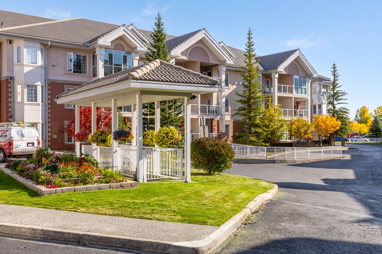 Multi-unit residential building with a covered entrance, flower beds, balconies, and autumn trees.