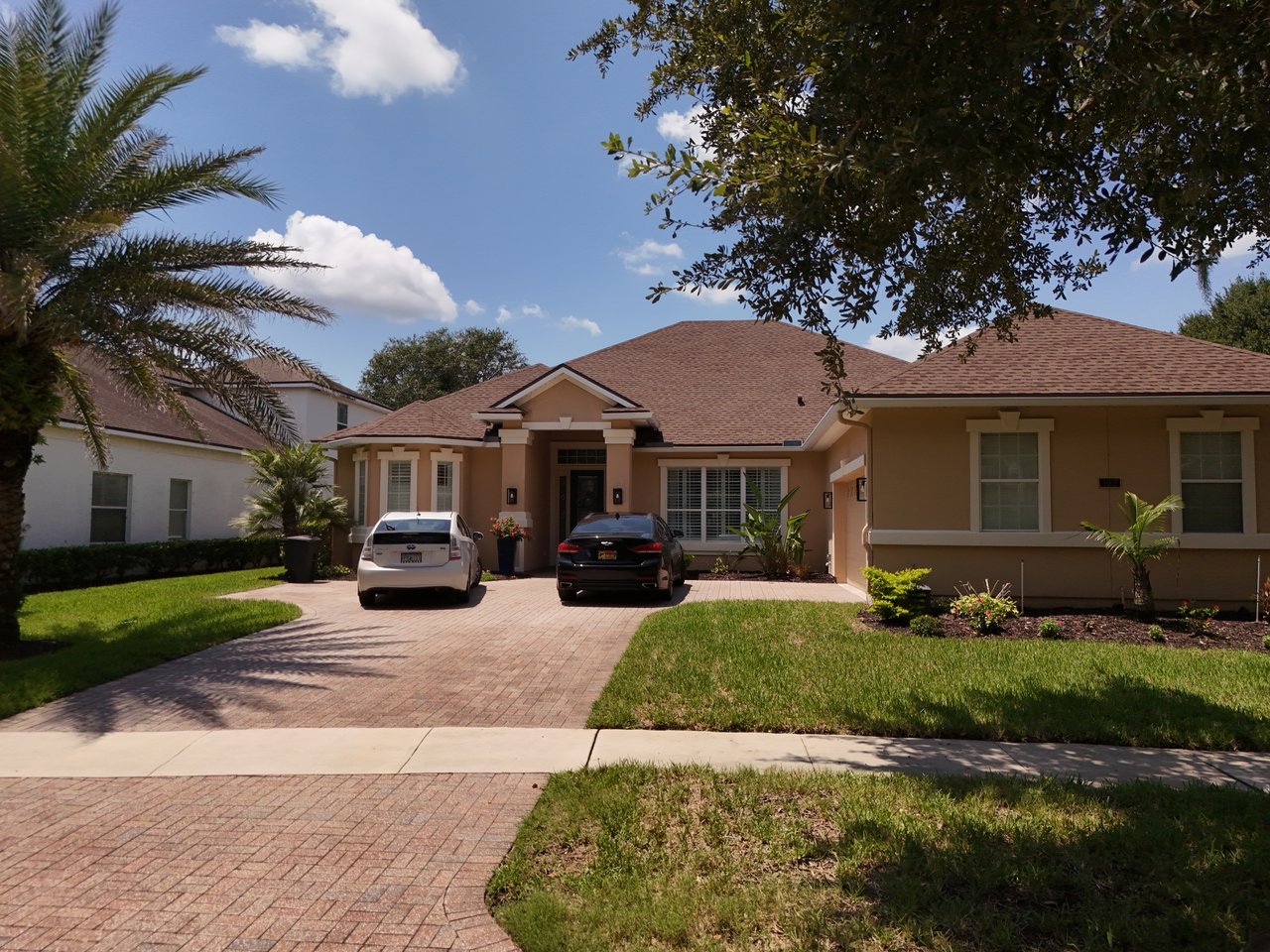 Tan single-story home with a brick driveway, parked cars, and a landscaped front lawn.