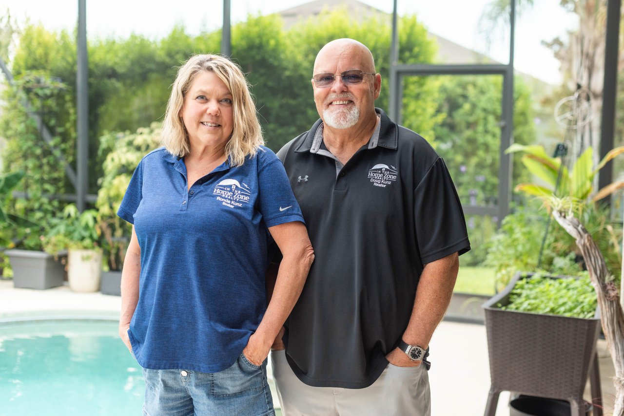 Two real estate agents pose beside a pool wearing In The Home Zone Realty shirts.