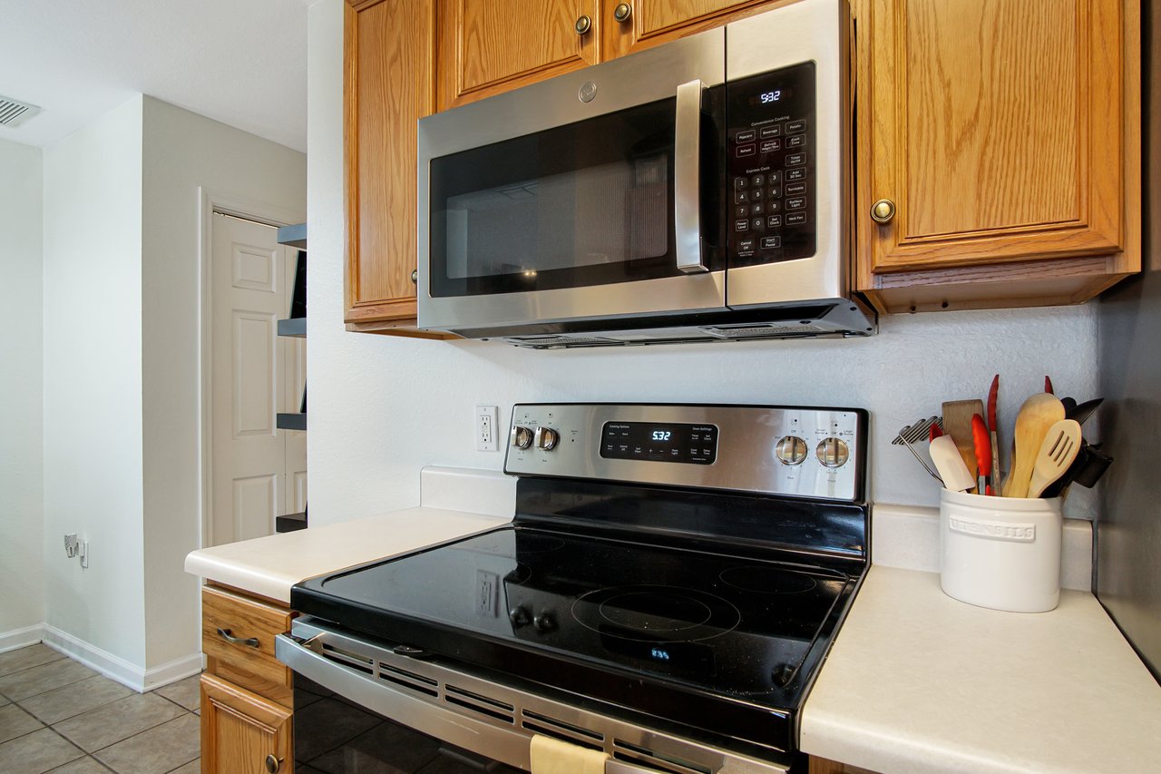 Modern kitchen with a stainless steel microwave and electric stove. Wooden cabinets above, utensils in a white holder on the right-side countertop.