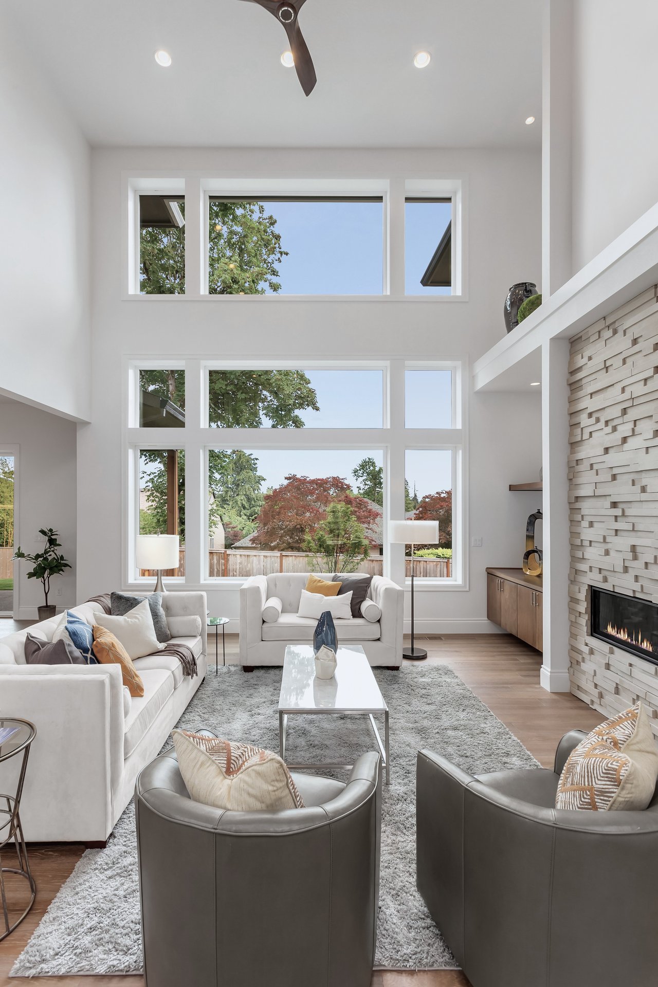 Double-height living room with tall windows, white sofas, and a stone fireplace.