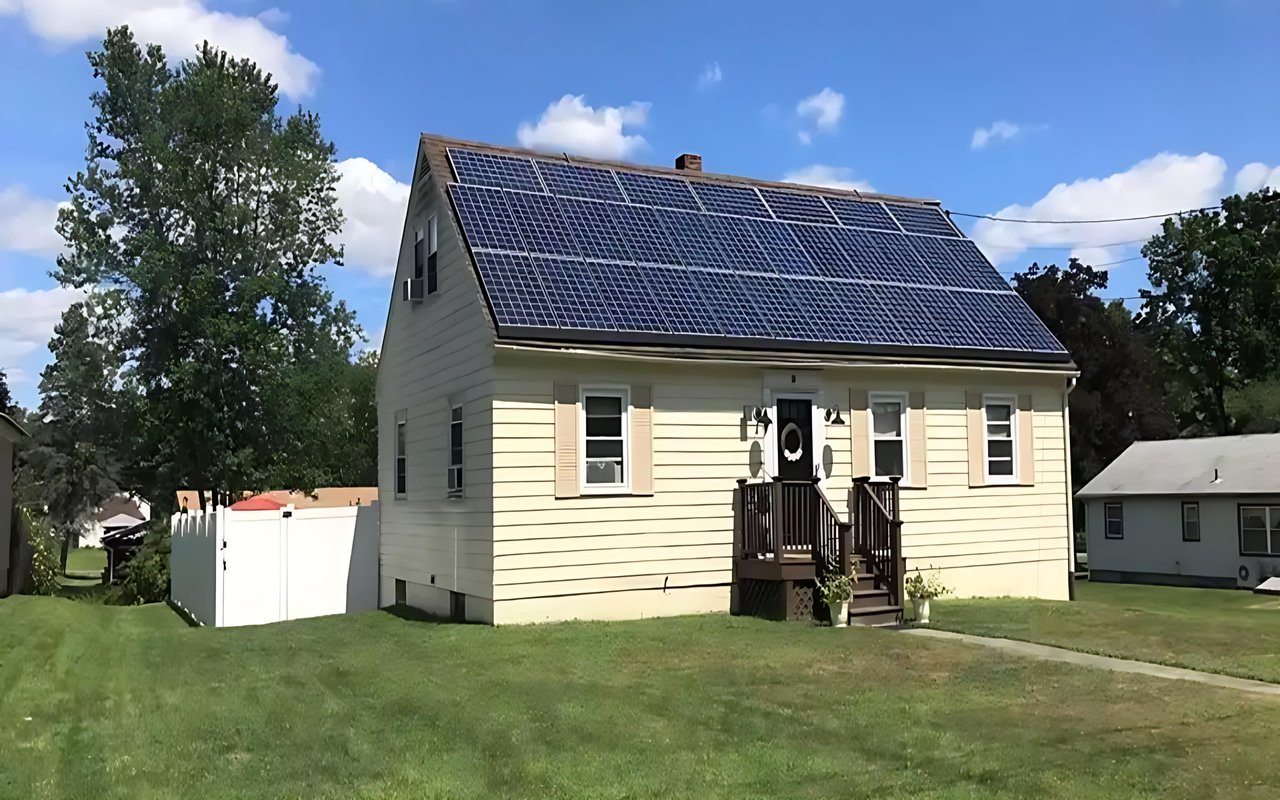 Pale-yellow house with roof-mounted solar panels, a small front porch, and a white shed beside the lawn.