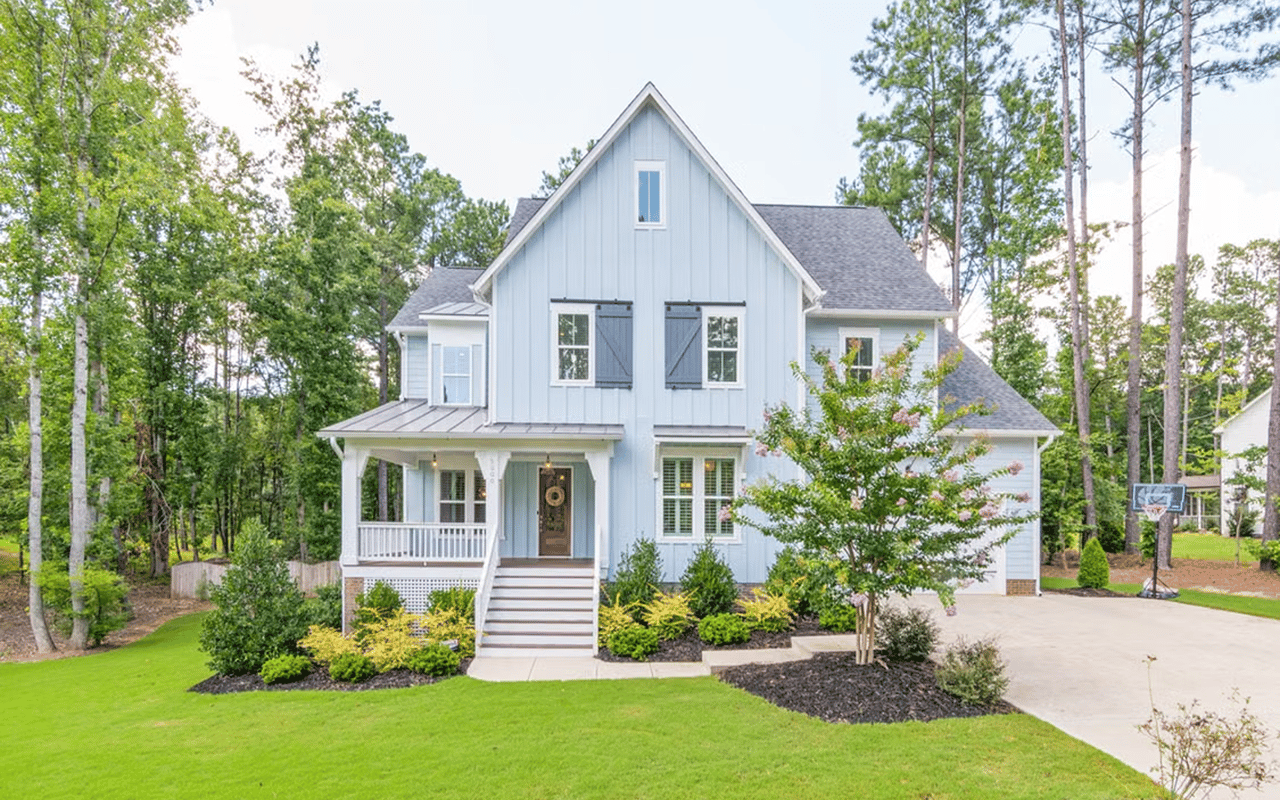 Light blue farmhouse with a covered porch, flowering landscaping, and a broad green lawn.