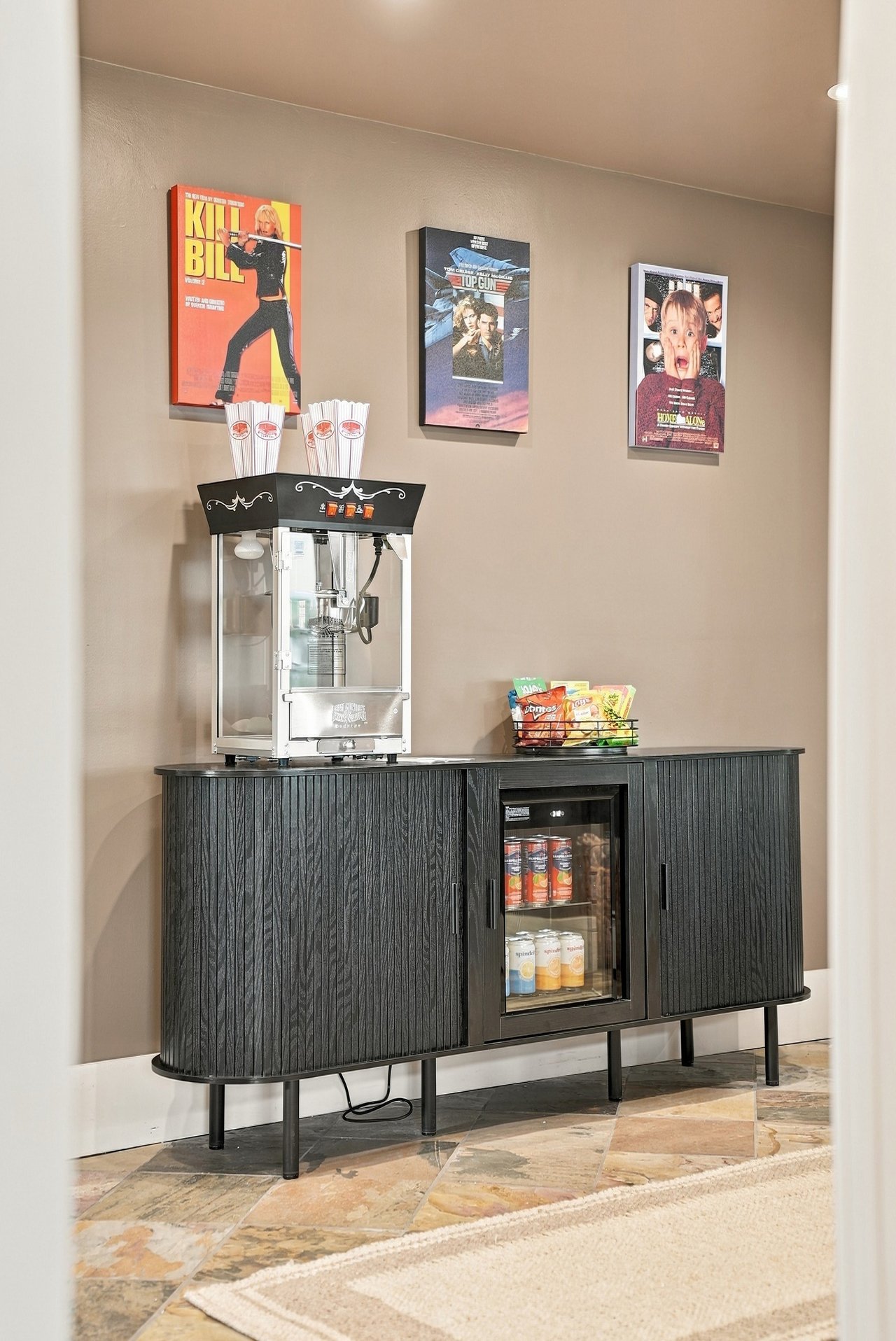 Media-room snack station with a popcorn machine, mini fridge, snacks, and framed movie posters.