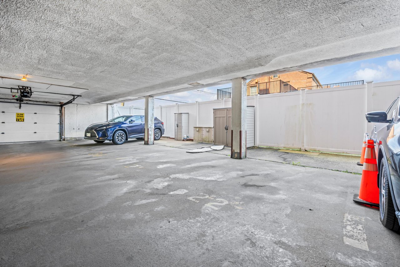 Covered parking garage with concrete support columns, parked cars, and storage cabinets along a white fence.