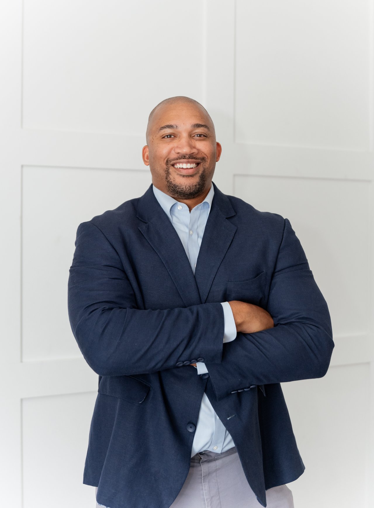 Wayne Whiting smiling with arms crossed in a navy suit against a white paneled wall.