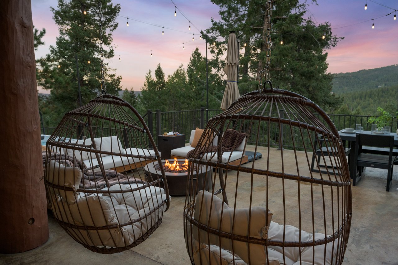 Patio with hanging egg chairs, a fire pit, string lights, and wooded hills at dusk.