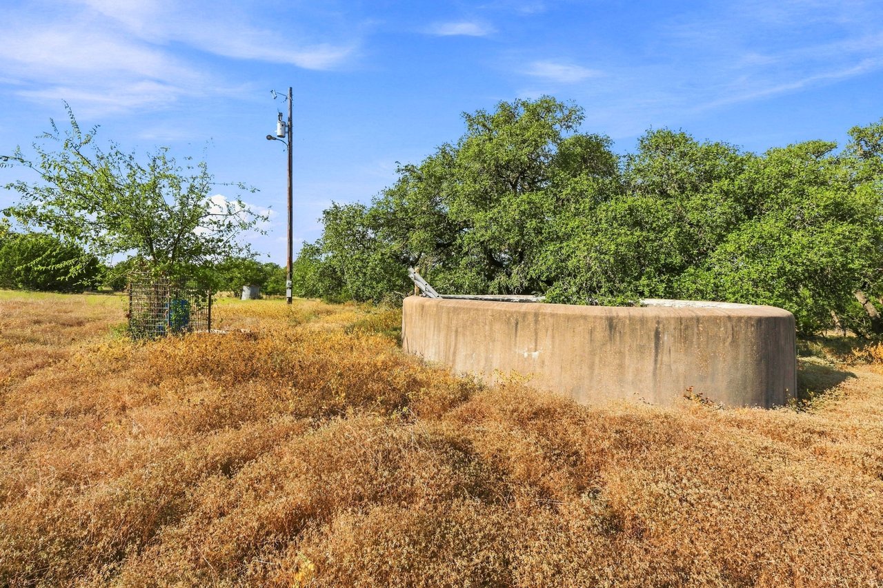 Ranch land with a large concrete water trough, dry grass, and scattered oak trees.