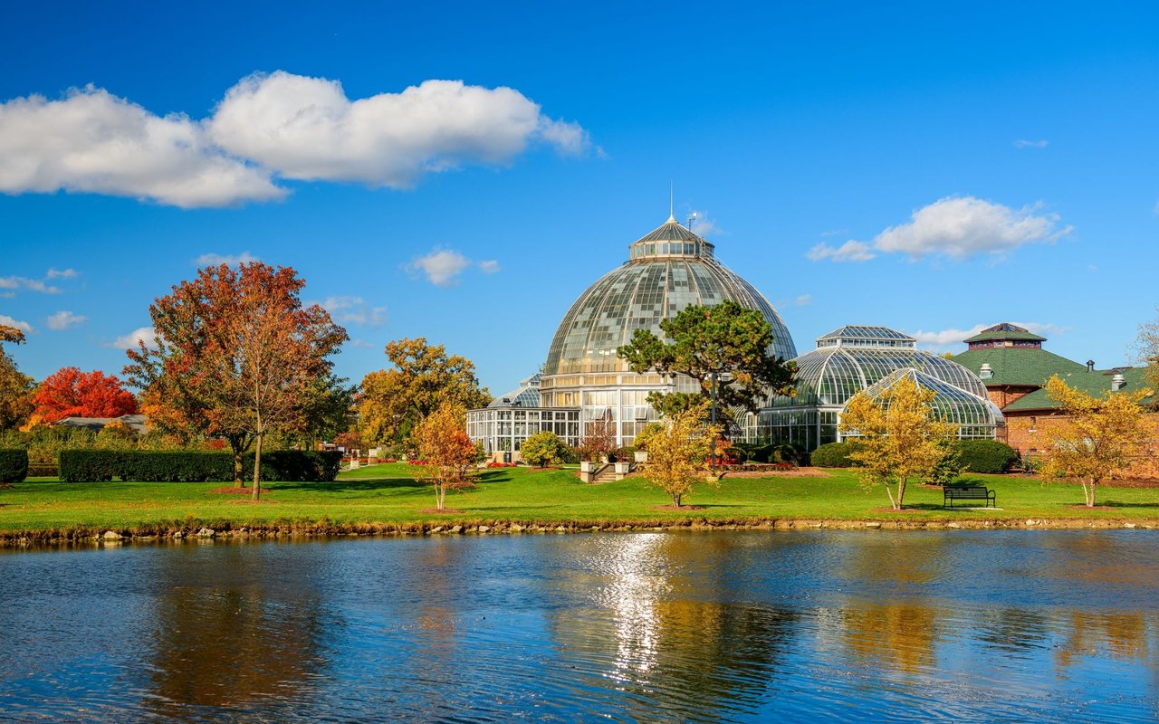 Glass-domed conservatory beside a pond, framed by autumn trees and green lawns.