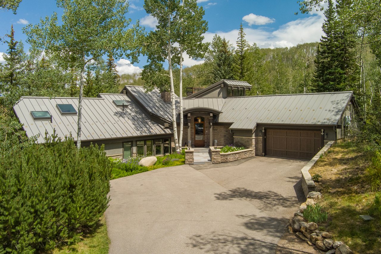 Modern mountain home with a broad metal roof, stone entry, and attached garage among mature trees.