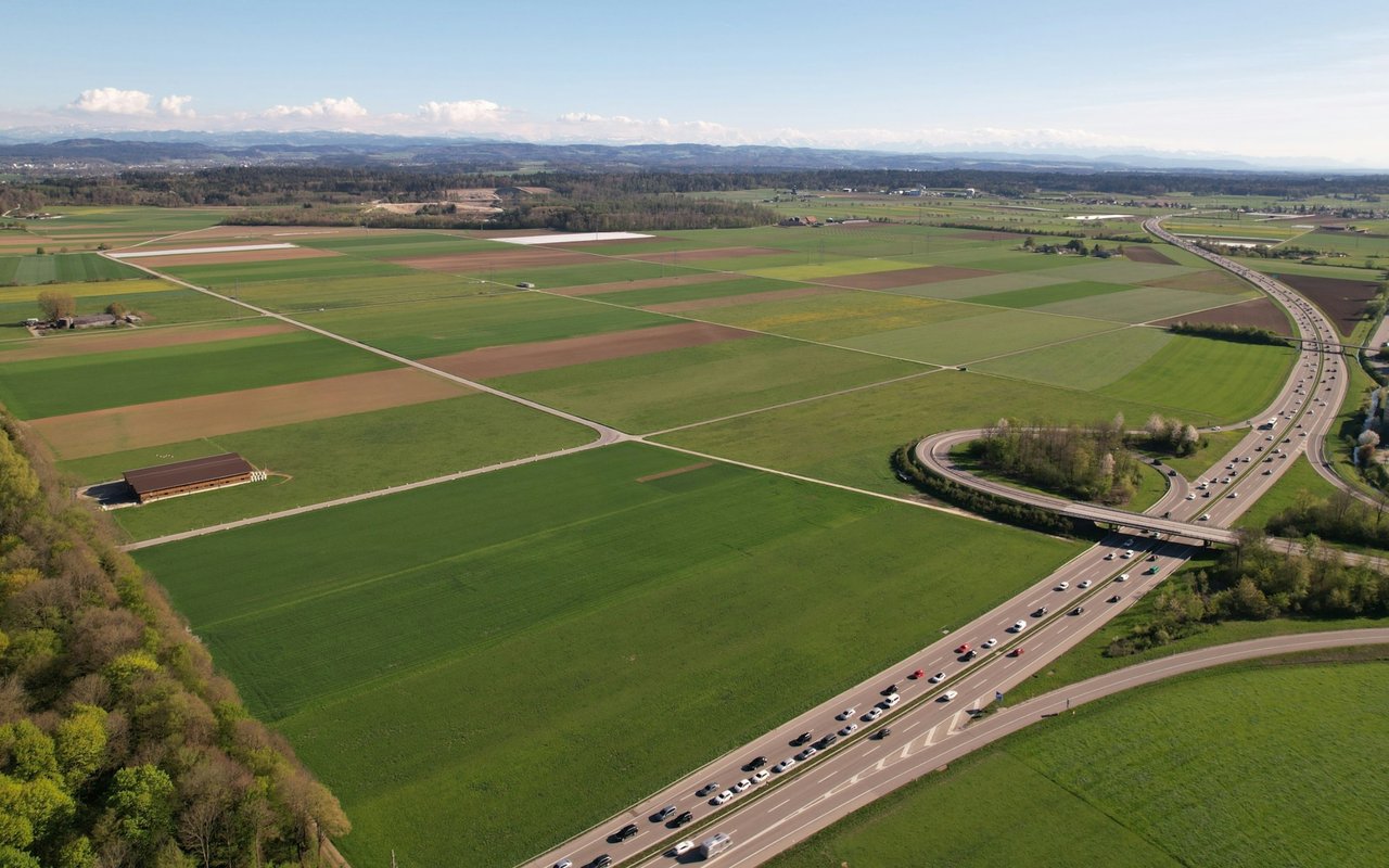Aerial view of patchwork farmland beside a busy highway interchange, with distant hills.