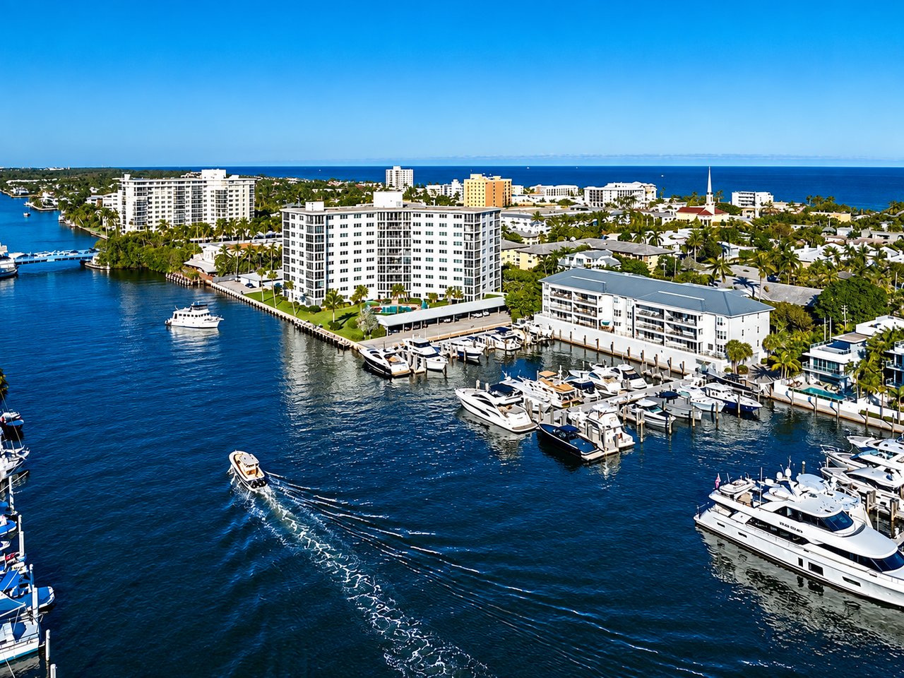 Aerial waterfront neighborhood with a marina, condominium buildings, and a boat crossing the channel.