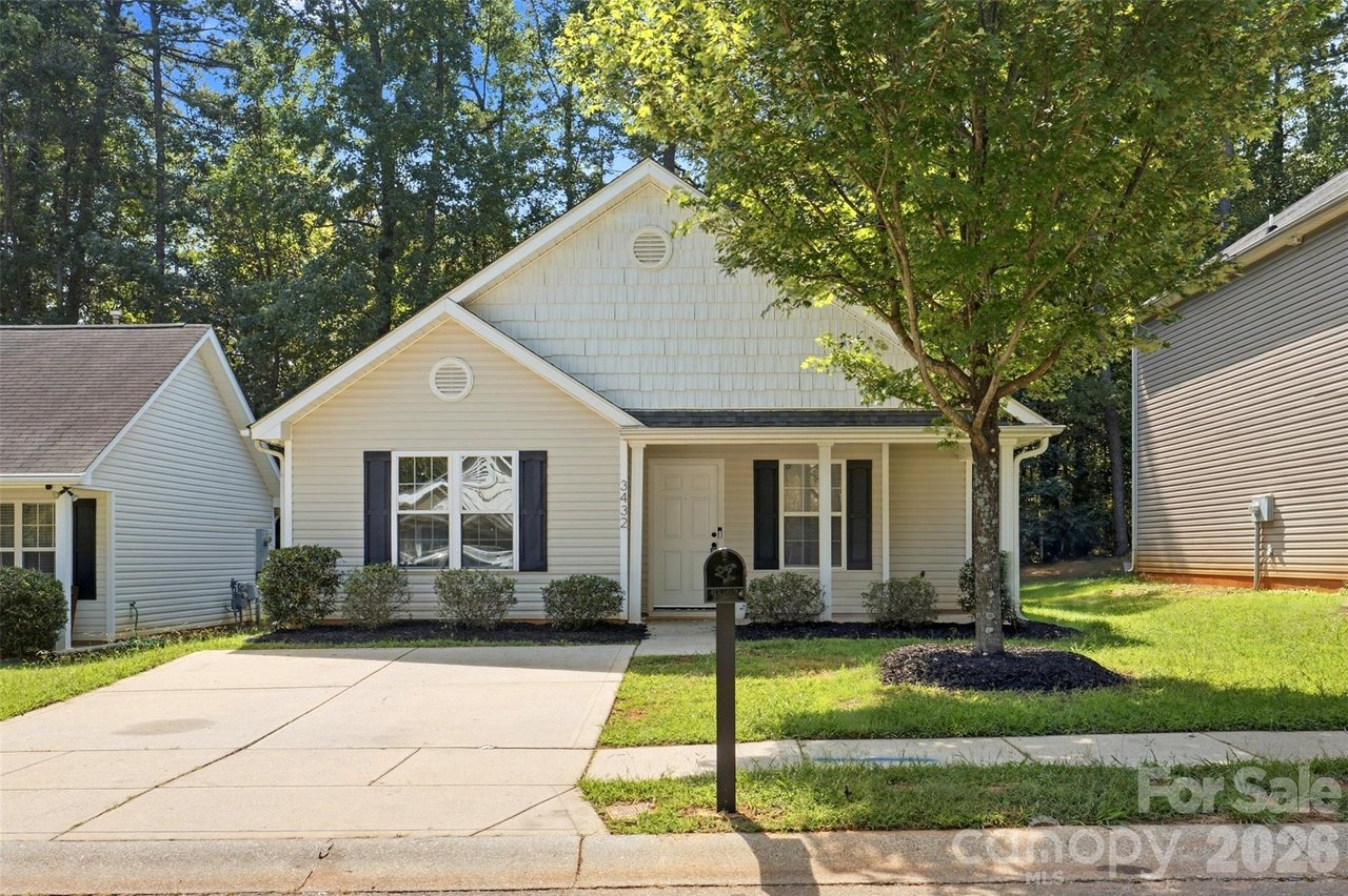 Light-sided house with a covered porch, dark shutters, and a driveway shaded by mature trees.