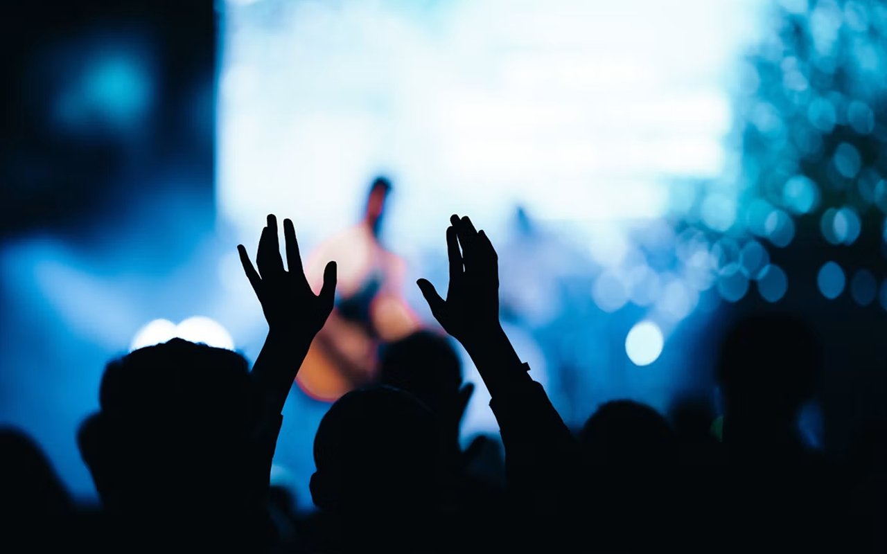 Concertgoers raise their hands toward a performer under blue stage lights.