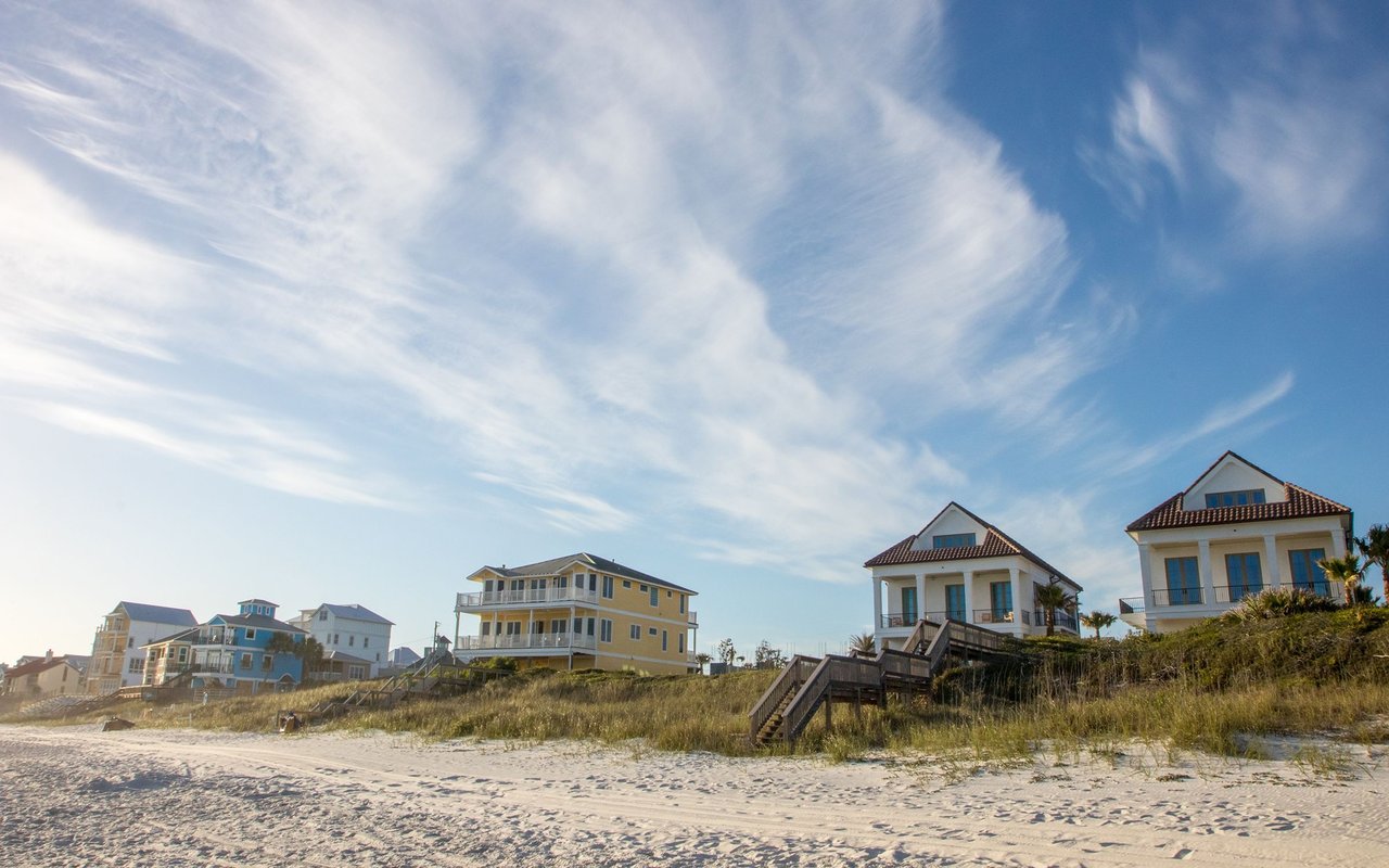 Coastal homes behind sandy dunes, with wooden boardwalk stairs leading to the beach.