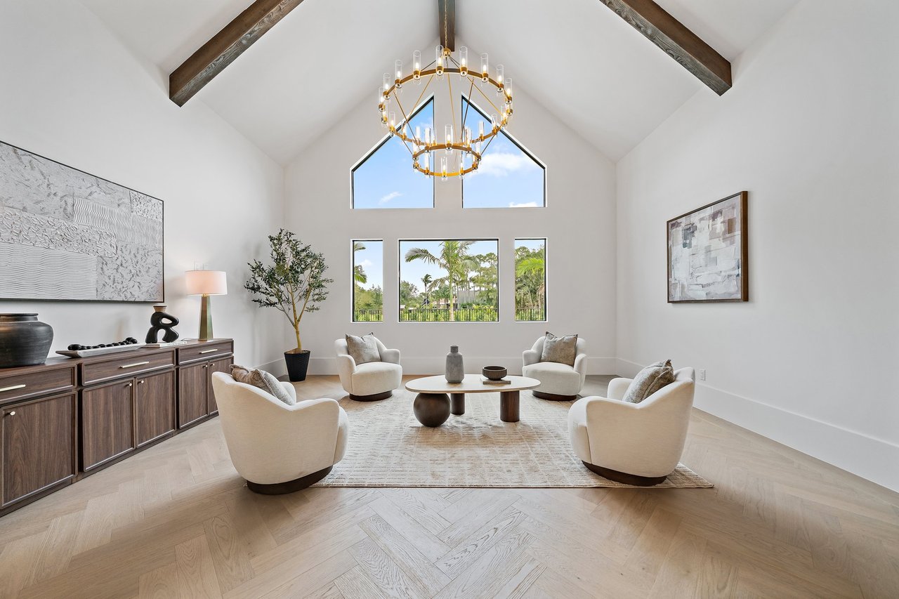 Vaulted living room with exposed wood beams, a gold chandelier, white lounge chairs, and palm views.