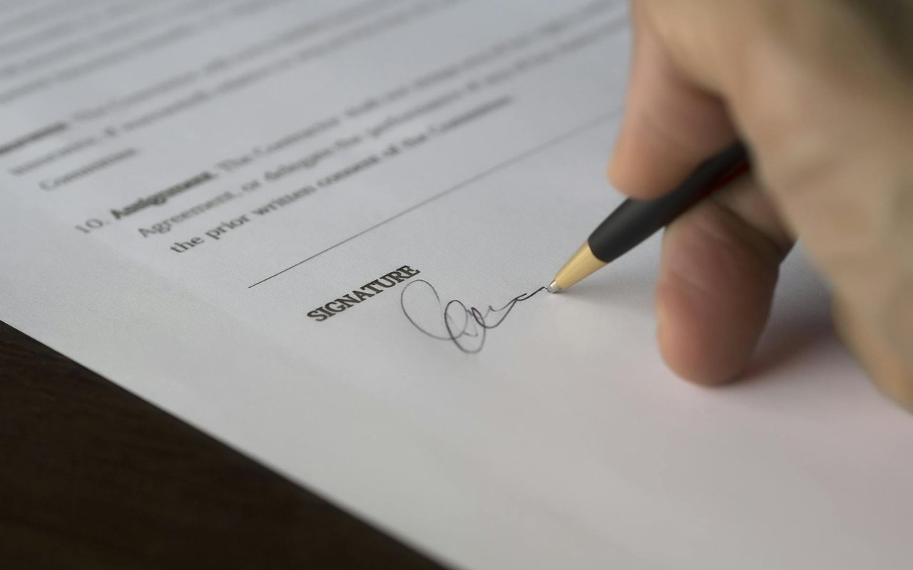 A hand signs a printed document beside the word “SIGNATURE.”