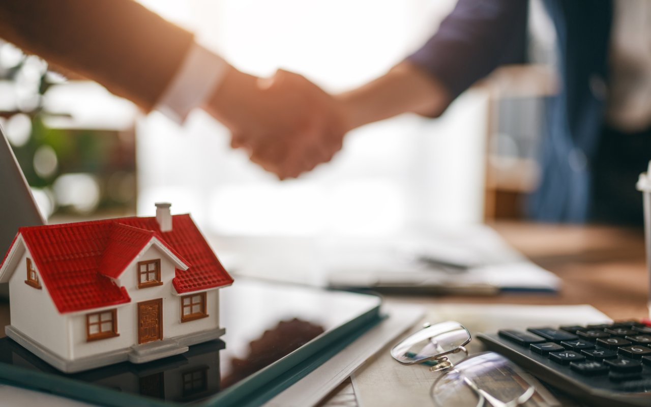 A miniature red-roofed house beside paperwork and a calculator as two people shake hands.