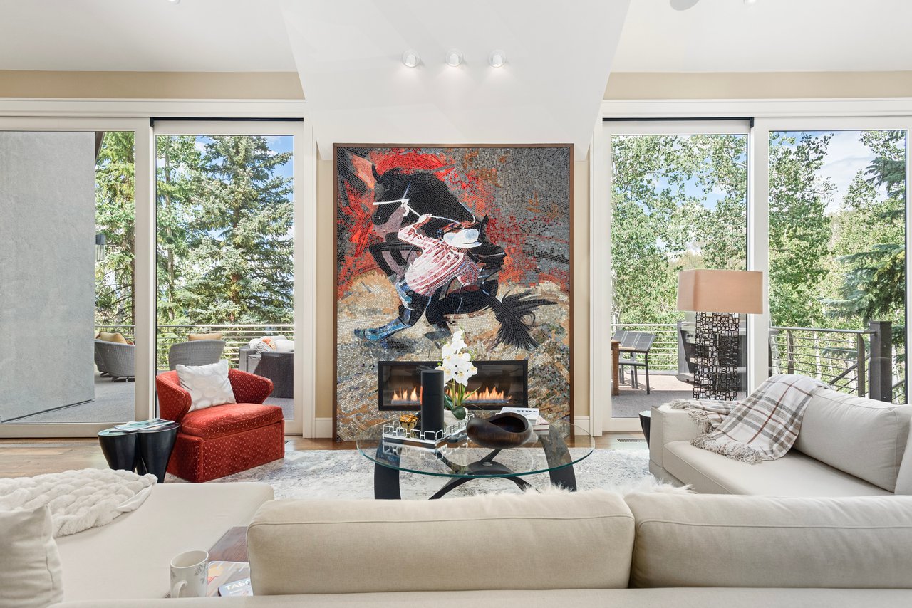 Living room with a mosaic fireplace, cream sofas, and glass doors opening onto a tree-lined deck.