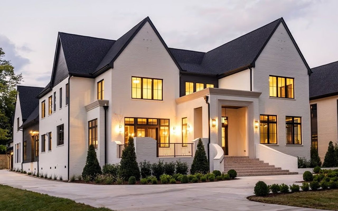 White-brick home with dark gabled roofs, glowing windows, broad entry steps, and landscaped grounds.