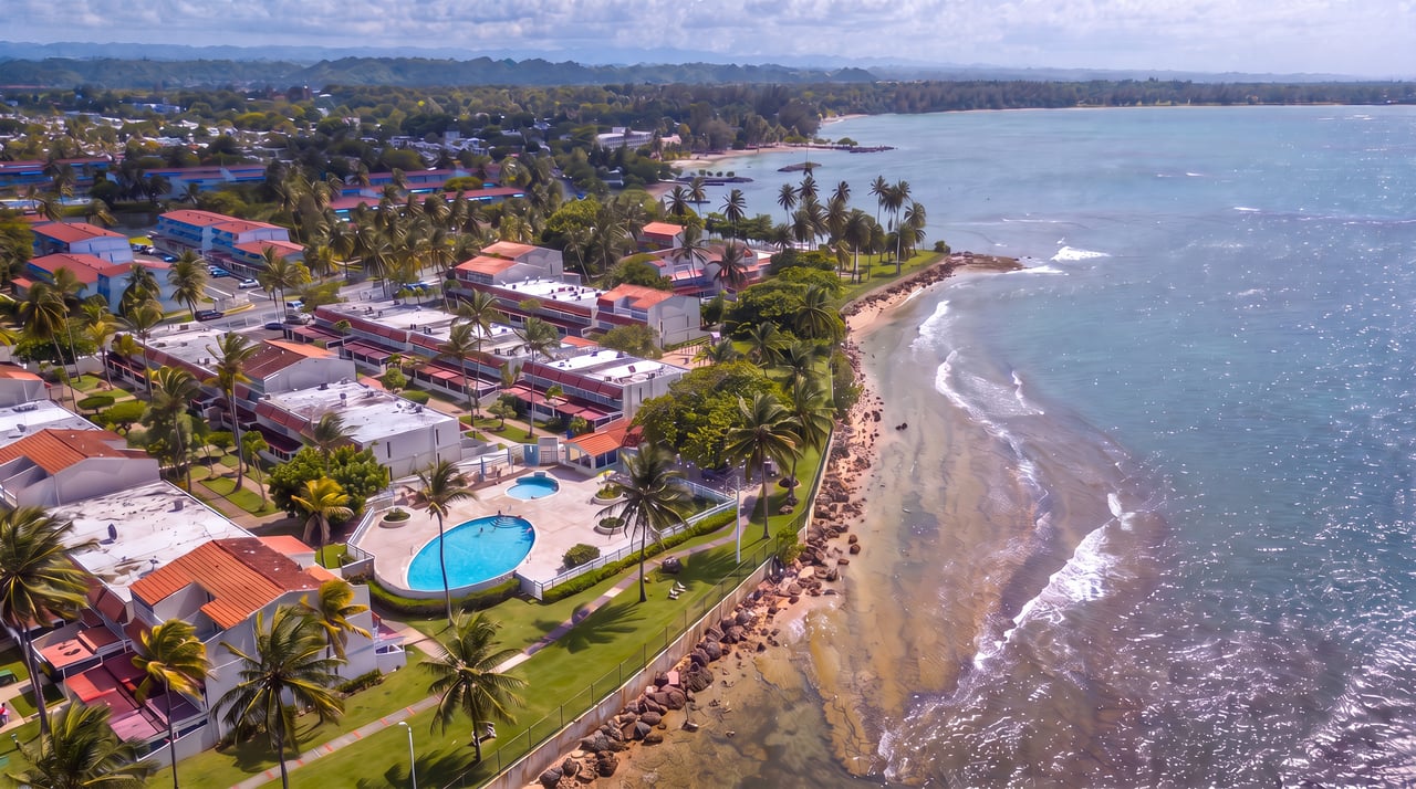Aerial view of beachfront villas, swimming pools, palm trees, and a sandy shoreline.