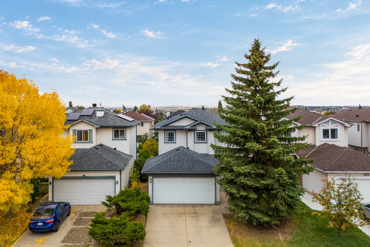Gray-sided house with a double garage, framed by a tall spruce and yellow autumn tree.
