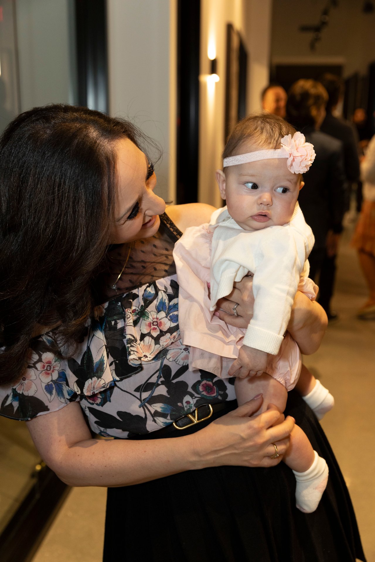 A woman in a floral blouse holds a baby wearing a pale pink flower headband.