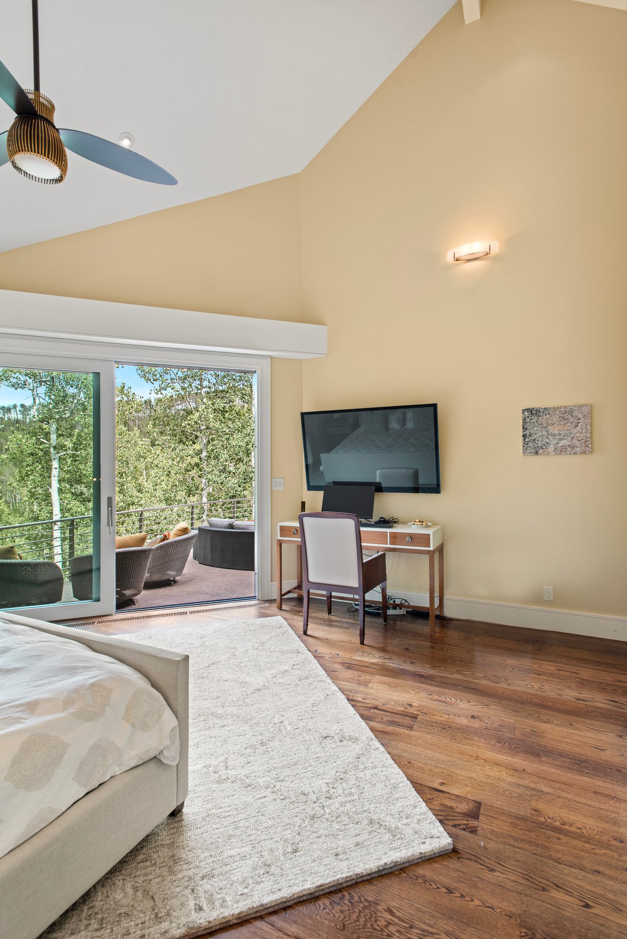 Furnished bedroom with sliding doors to a tree-lined balcony and a desk beneath a wall-mounted television.