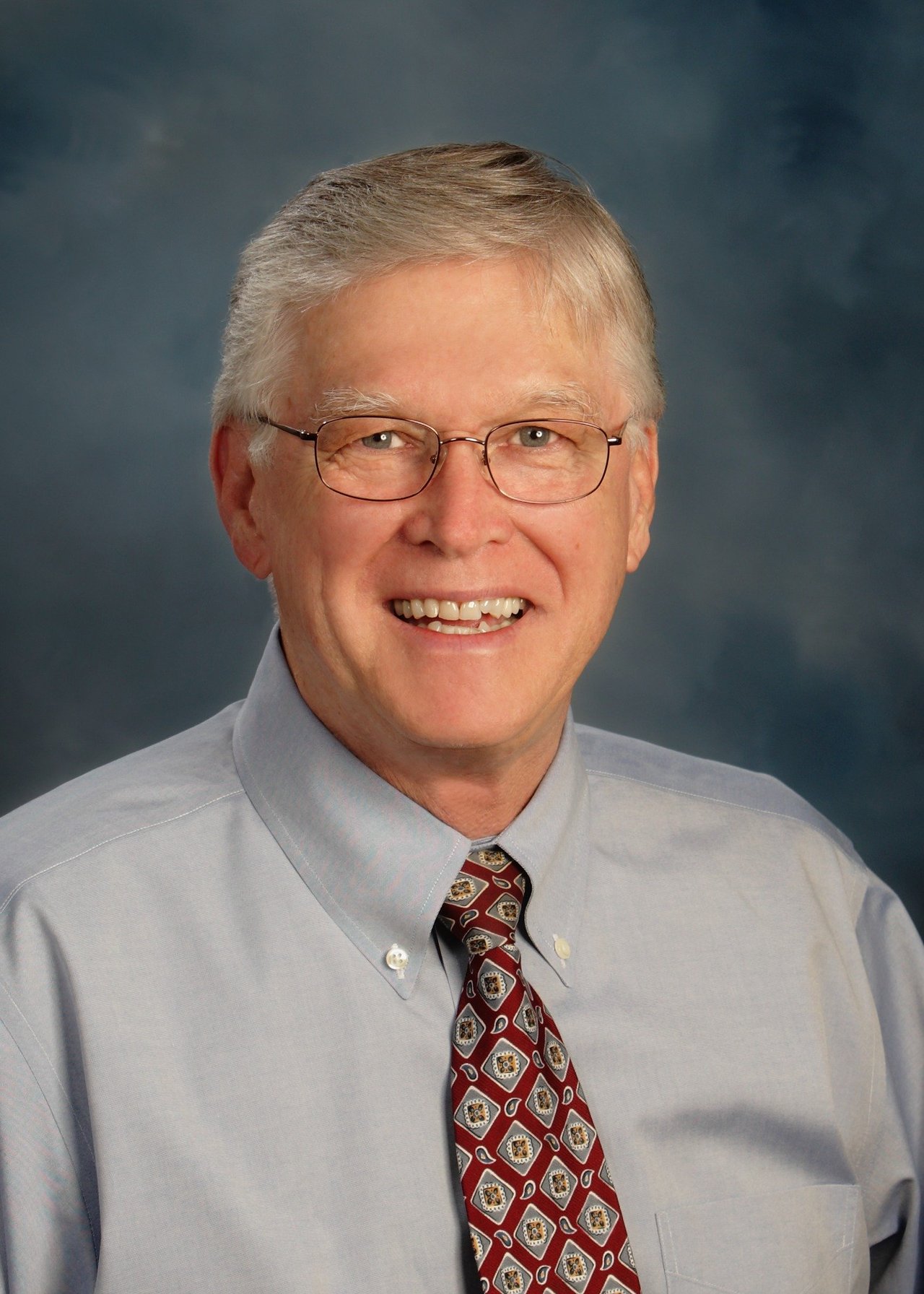 Dan Goad smiling in glasses, a light blue shirt, and patterned tie.