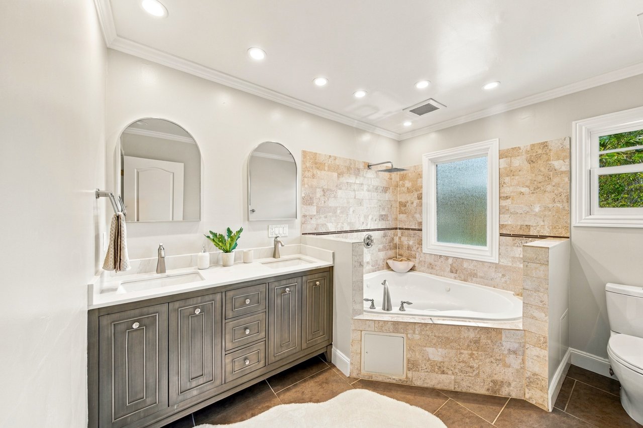 Bathroom with a double-sink gray vanity and a tiled corner bathtub beneath a frosted window.