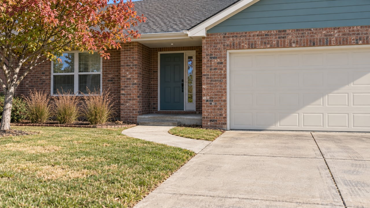 Red-brown brick ranch with a teal entry door, cream garage door, concrete approach and an autumn lawn.