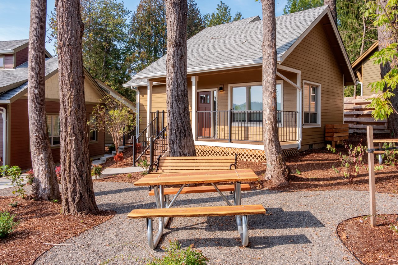 Tan home with a covered porch, framed by tall trees and a wooden picnic table.