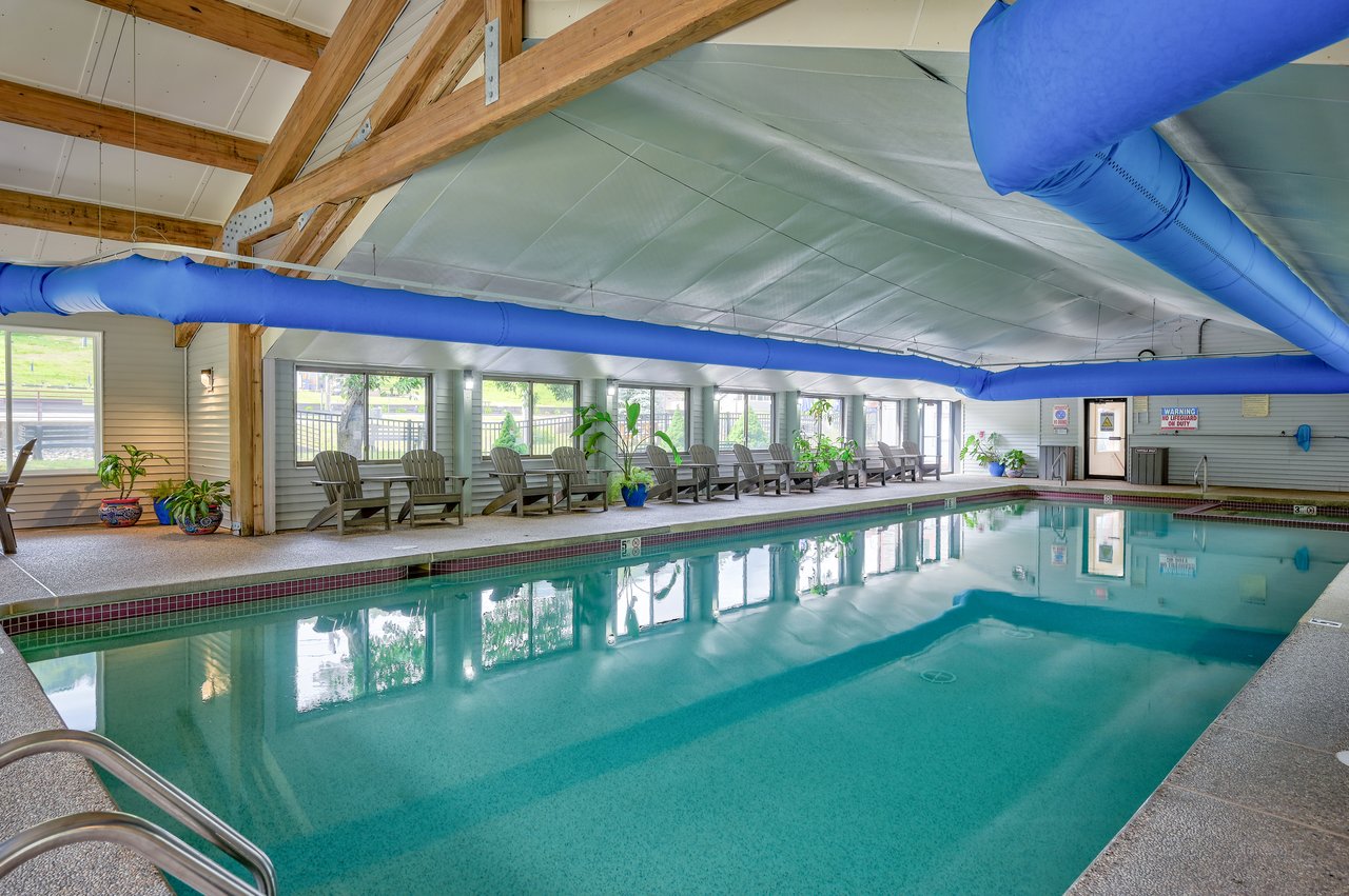 Indoor swimming pool with exposed timber beams, blue ducts, and lounge chairs along the windows.