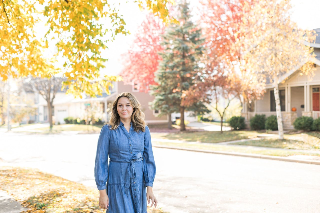 A prospective first-time homebuyer in a blue dress walking down a quiet suburban street lined with single-family homes and colorful autumn trees.