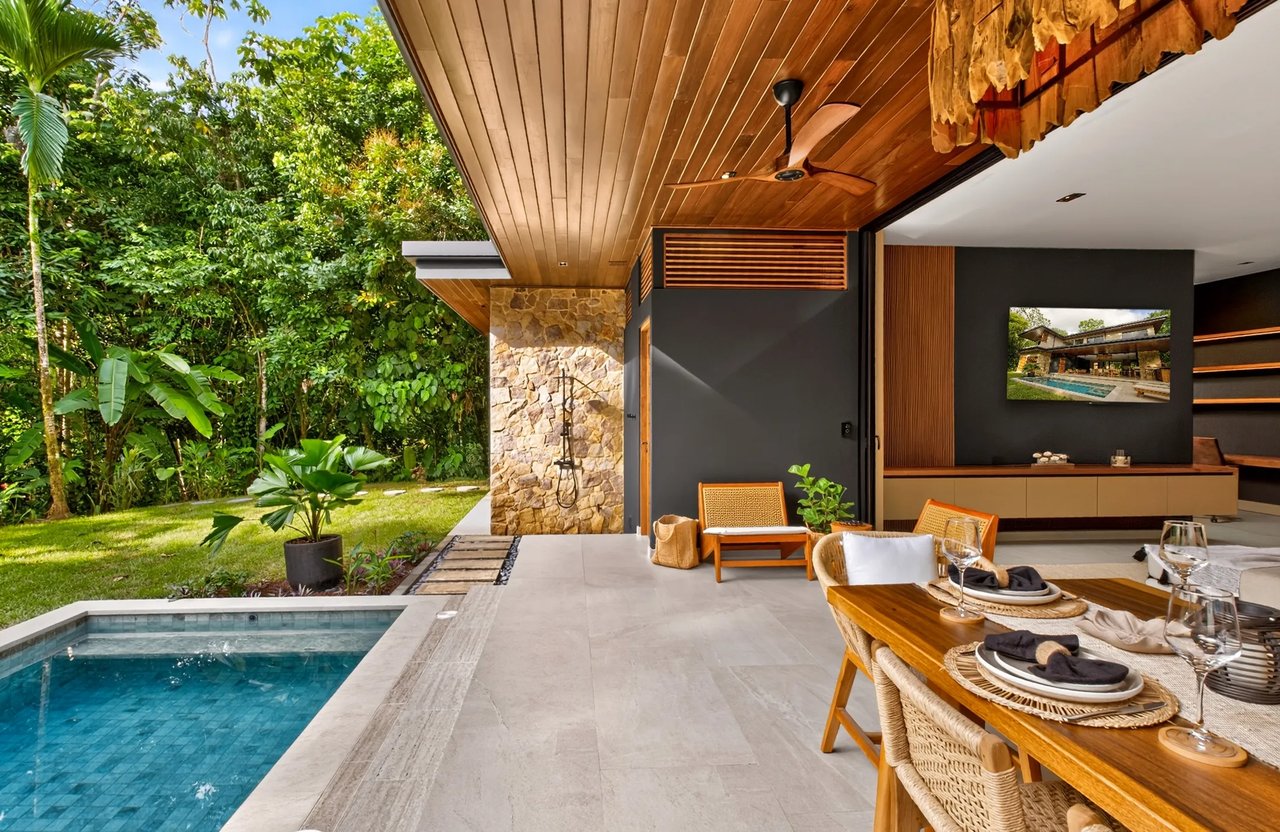 Covered patio with a dining table and lounge chairs beside a pool, surrounded by tropical greenery.